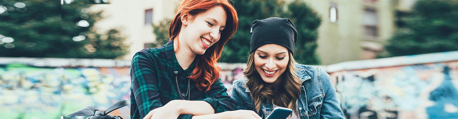 Programa Joven In - Dos chicas jóvenes sonriendo mientras están con el móvil en la mano sentadas en un parque