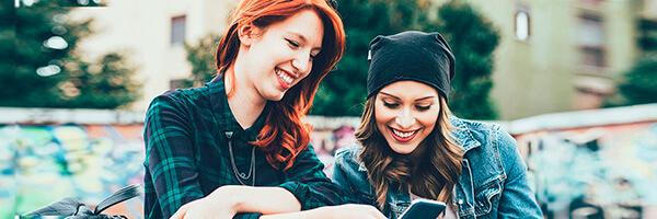 Programa Joven In - Dos chicas jovenes sonriendo mientras navegan por intenet con el movil en un parque